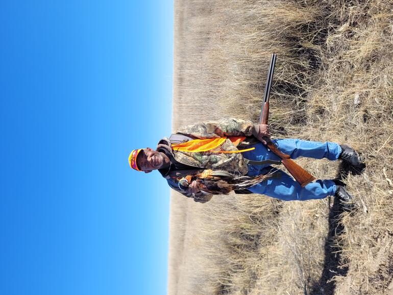 A person wearing camouflage and blaze orange shands in a field holding a pheasant and a shotgun under a clear blue sky.