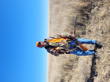 A person wearing camouflage and blaze orange shands in a field holding a pheasant and a shotgun under a clear blue sky.