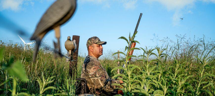 A man in camouflage stands in tall grass next to a dove decoy, blending into the natural surroundings