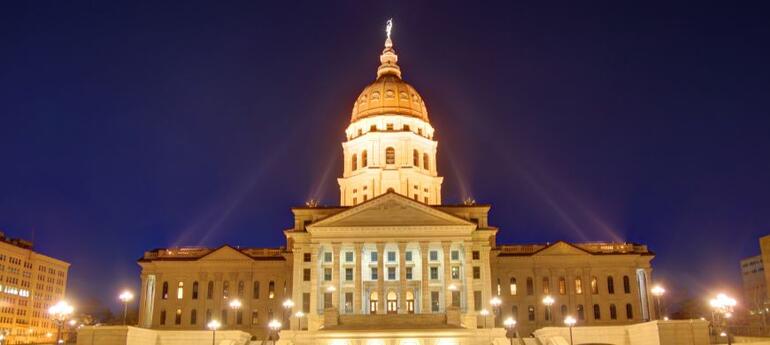 Illuminated Topeka Capitol building with a prominent dome at night, featuring classical architecture and columns, surrounded by streetlights.